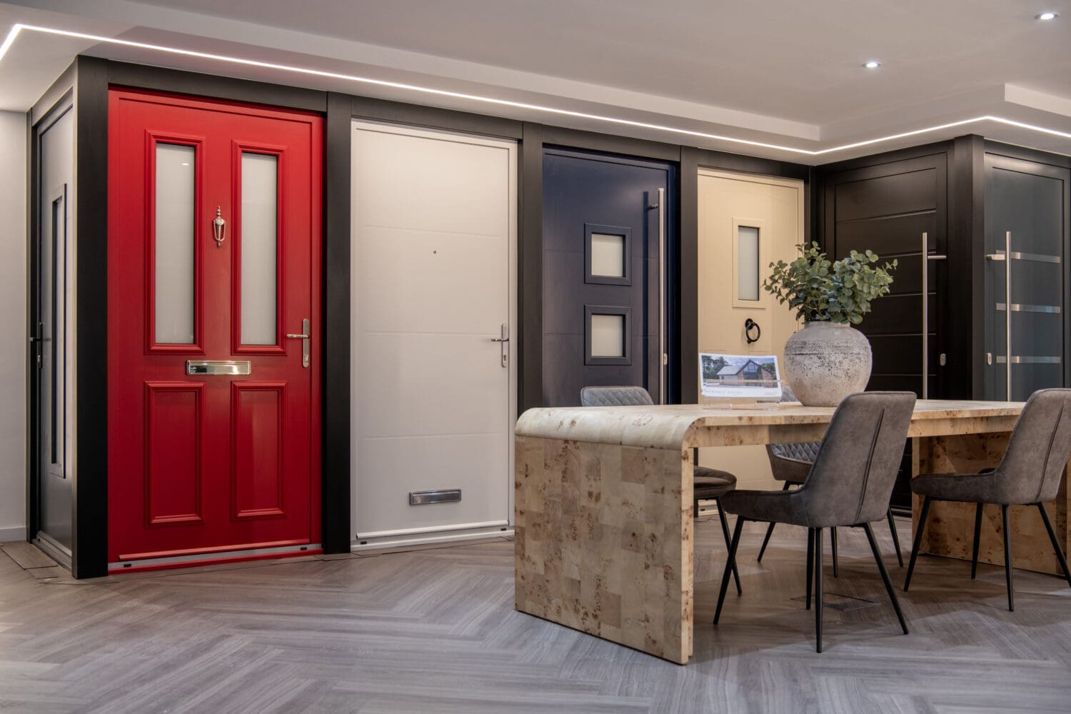 A showroom displays various front doors in different colors and styles, including a red door, beneath sleek glass roofing, behind a modern wooden table with chairs and a large vase with green foliage.