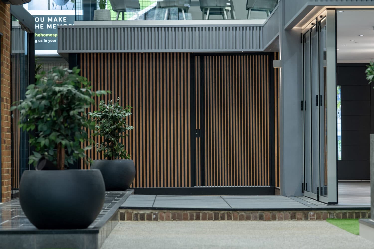 A modern indoor space with large black planters holding green plants, a bench, and a wooden slat wall in the background. The area is well-lit with natural light from bespoke glass solutions above.