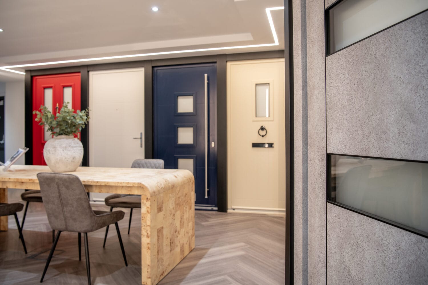 A modern showroom with four colorful front doors (red, white, blue, and beige) displayed behind a light wood table with grey chairs and a large vase of greenery. The space features herringbone flooring and glass roofing for natural light.