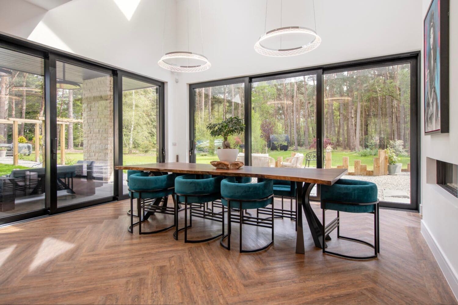 A modern dining room with a large wooden table, teal velvet chairs, and floor-to-ceiling bifold doors offering views of a lush garden and forest. The space features wood flooring and two circular pendant lights overhead.
