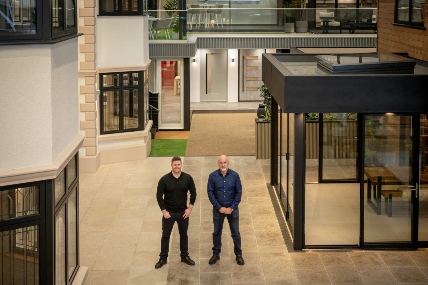 Two men stand side by side in a modern, indoor courtyard with large windows, tiled flooring, and sleek glass roofing highlighting the clean, contemporary space with its mix of glass and brick accents.