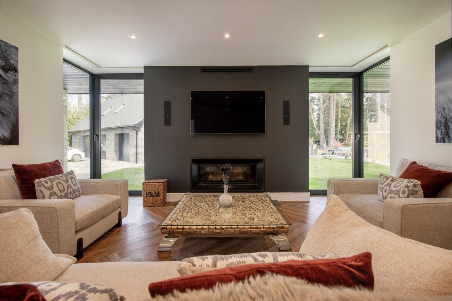 Modern living room with neutral-toned sofas, patterned cushions, a coffee table, and a dark accent wall featuring a mounted TV above a fireplace. Large sliding doors open to reveal a green outdoor area.