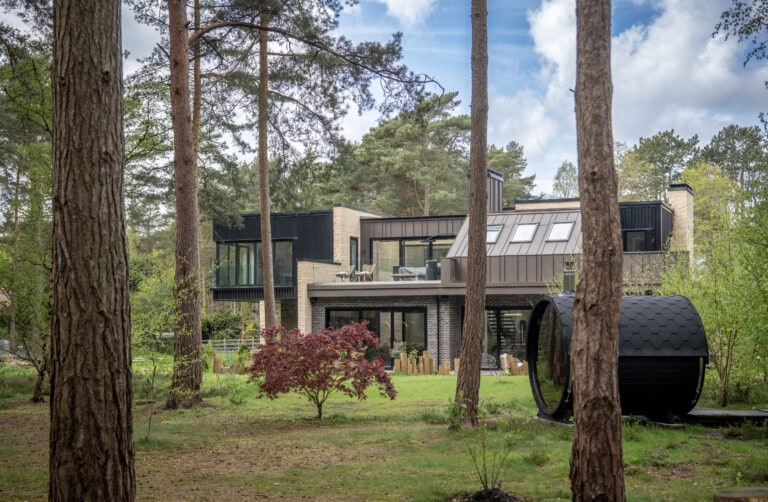 Modern two-story house with large windows featuring bespoke glass solutions and balconies, surrounded by tall trees and greenery. A small red-leafed tree is in the yard, near a round black outdoor structure. The setting is peaceful and wooded.