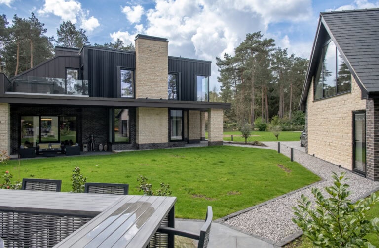 Modern two-story house with stone and dark accents, large windows, sliding doors, a balcony, and a detached garage. A patio with an outdoor table and green lawn are in the foreground, surrounded by trees and blue sky with clouds.