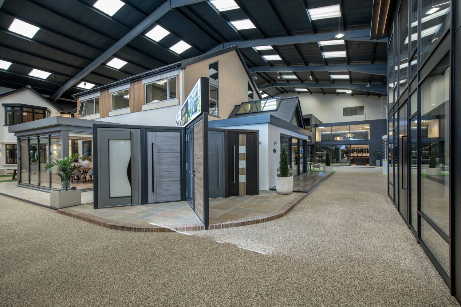 Modern indoor showroom featuring full-scale house models and a variety of exterior and bifold doors on display, with ample lighting from overhead skylights and large windows, plus clean walkways throughout the space.