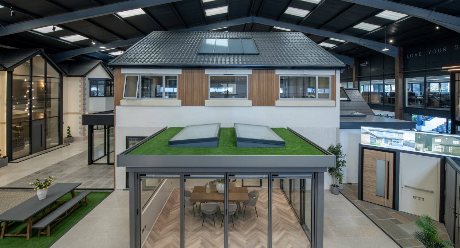 Modern showroom interior displaying a two-story house model with a glass-walled extension featuring bifold doors to the dining area, artificial grass roof, and skylights; other model homes visible in the background.