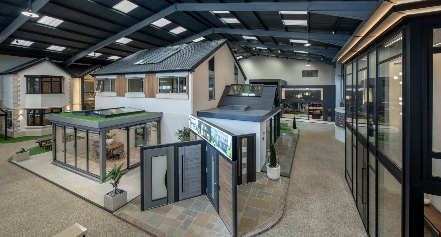 A spacious indoor showroom displays several modern house models with large windows, bespoke glass solutions for front doors, and outdoor patio areas, all under a high ceiling with skylights.