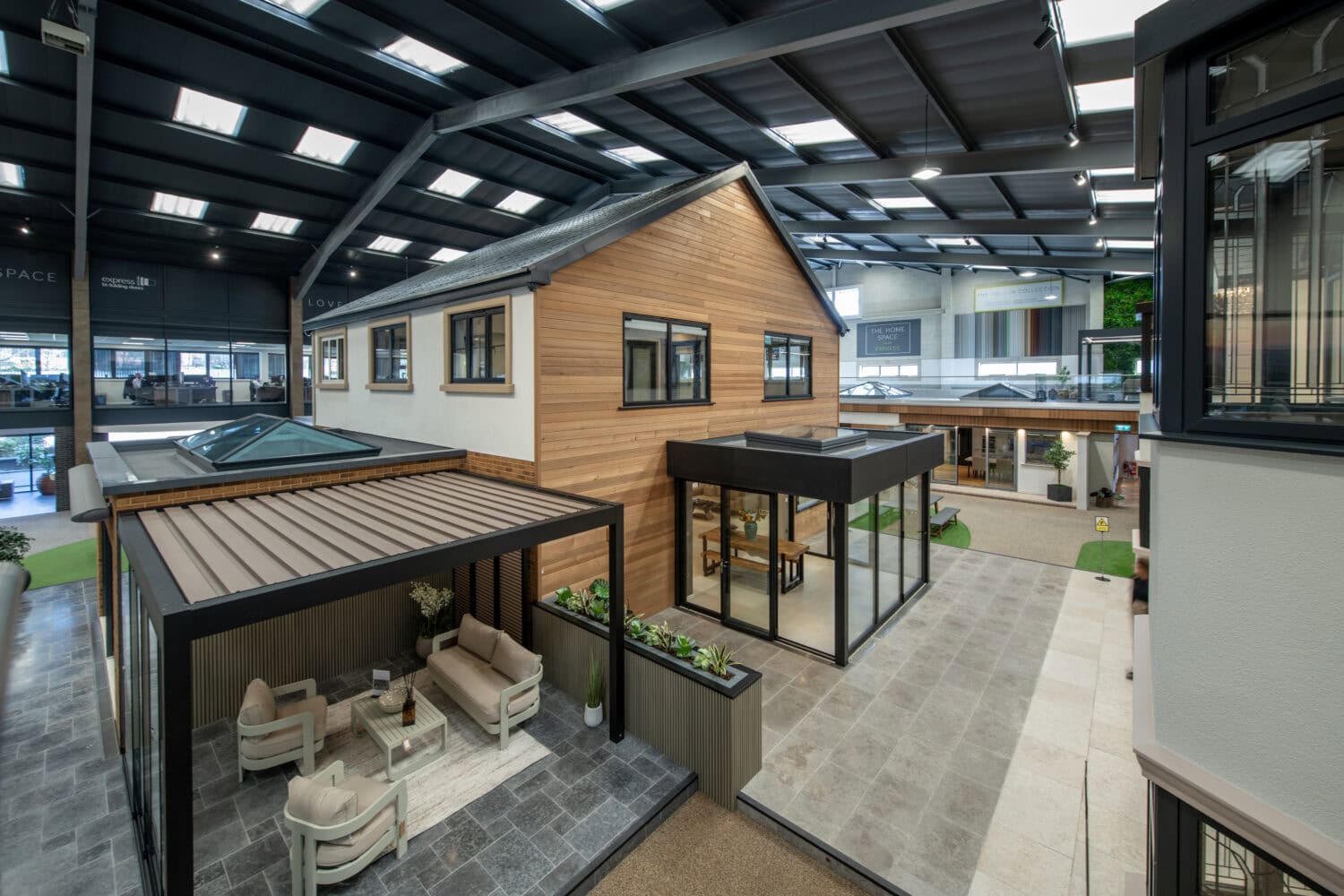 Indoor showroom featuring a modern two-story house with wood and white exterior, large windows, sleek bifold doors opening to a glass-enclosed patio, and an outdoor seating area with cushioned chairs and a sofa under a pergola.