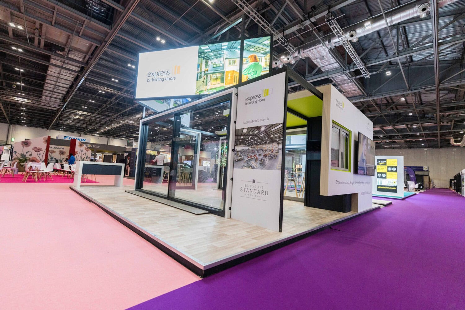 A modern exhibition booth with large glass sliding doors and bright signage for Express Bi-folding Doors stands on a wooden platform, showcasing stylish bifold doors in a spacious, well-lit event hall with a pink carpet.