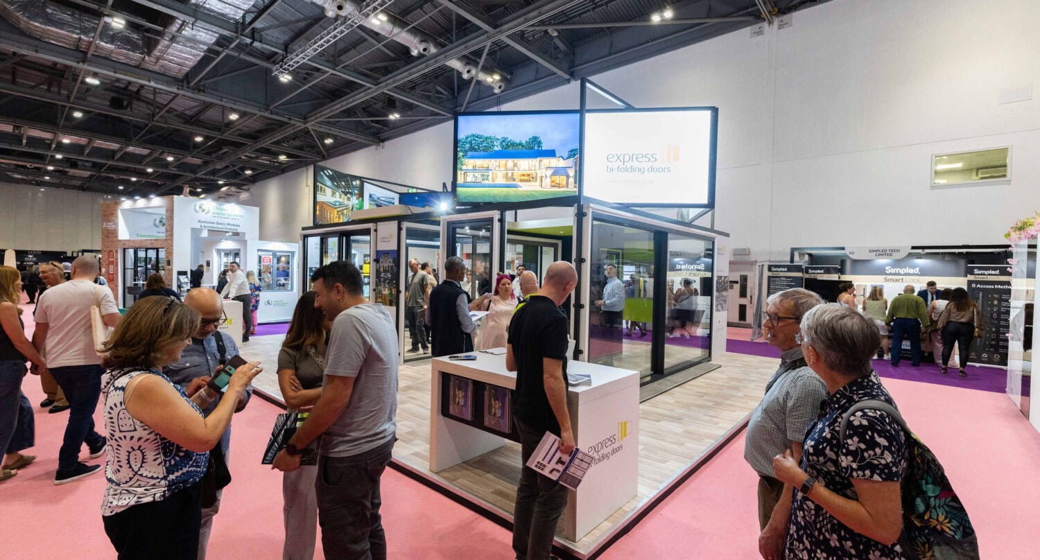 People walk around and interact at a busy indoor trade show, with various booths featuring bifold doors and glass displays under bright lighting. A large sign reads Express Bi-Folding Doors above one of the exhibits.