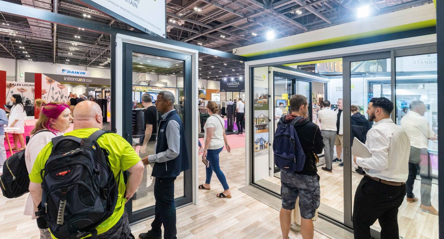 A group of people converse and browse at a busy trade show or exhibition hall, surrounded by displays and booths featuring large windows, informational signs, and impressive glass roofing overhead.