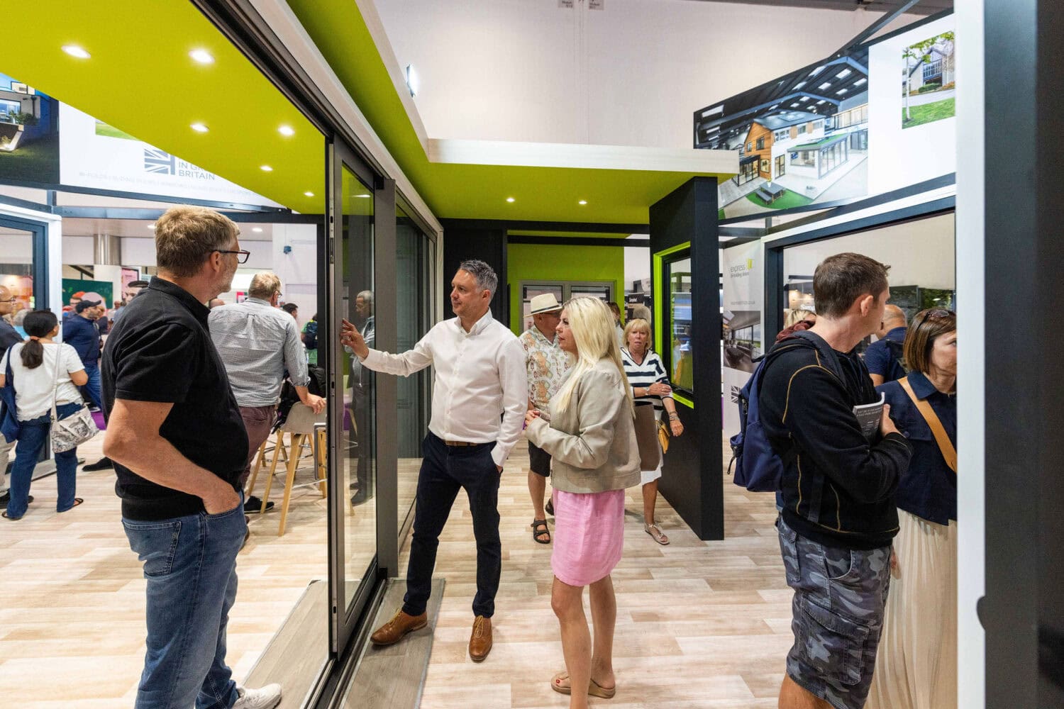 People interact and examine features in a modern showroom with bespoke glass solutions, including large glass doors and green accents. Some stand and talk while others look around; informational displays and photos are visible in the background.