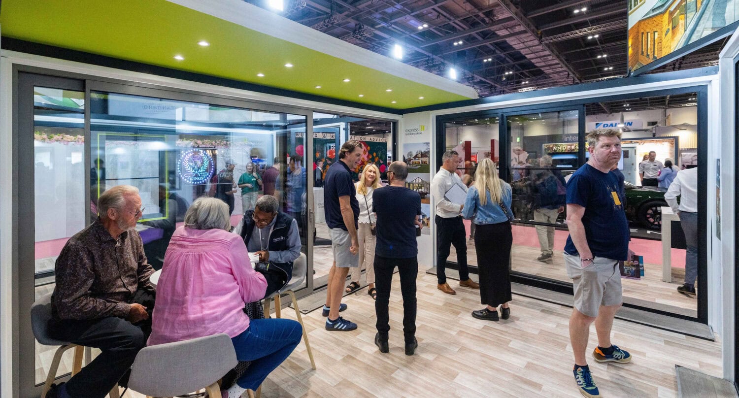 A group of people socializes and converses inside and around a modern indoor booth with sliding doors at a trade show or exhibition, featuring colorful displays and bright lighting in the background.