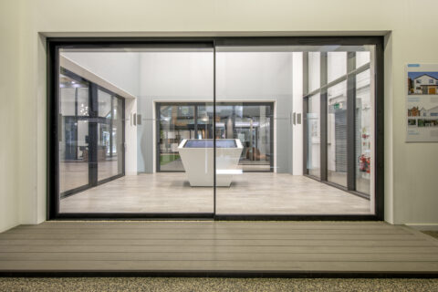 Large glass front doors open to a modern, minimalist interior with light wood flooring. Inside, a white, angular reception desk stands under bright lighting. A poster about houses is displayed on the right wall.