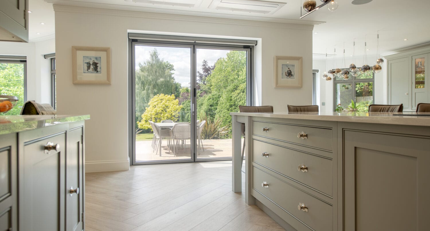 Modern kitchen with light gray cabinets and large island, featuring sliding glass doors that open to a patio with a table and chairs, overlooking a lush green garden. Sunlight brightens the space.
