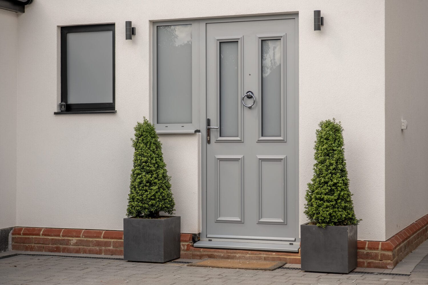A modern light gray front door with frosted glass panels, flanked by two tall, cone-shaped evergreen shrubs in square planters. The door is set in a white exterior wall with a small window to the left.