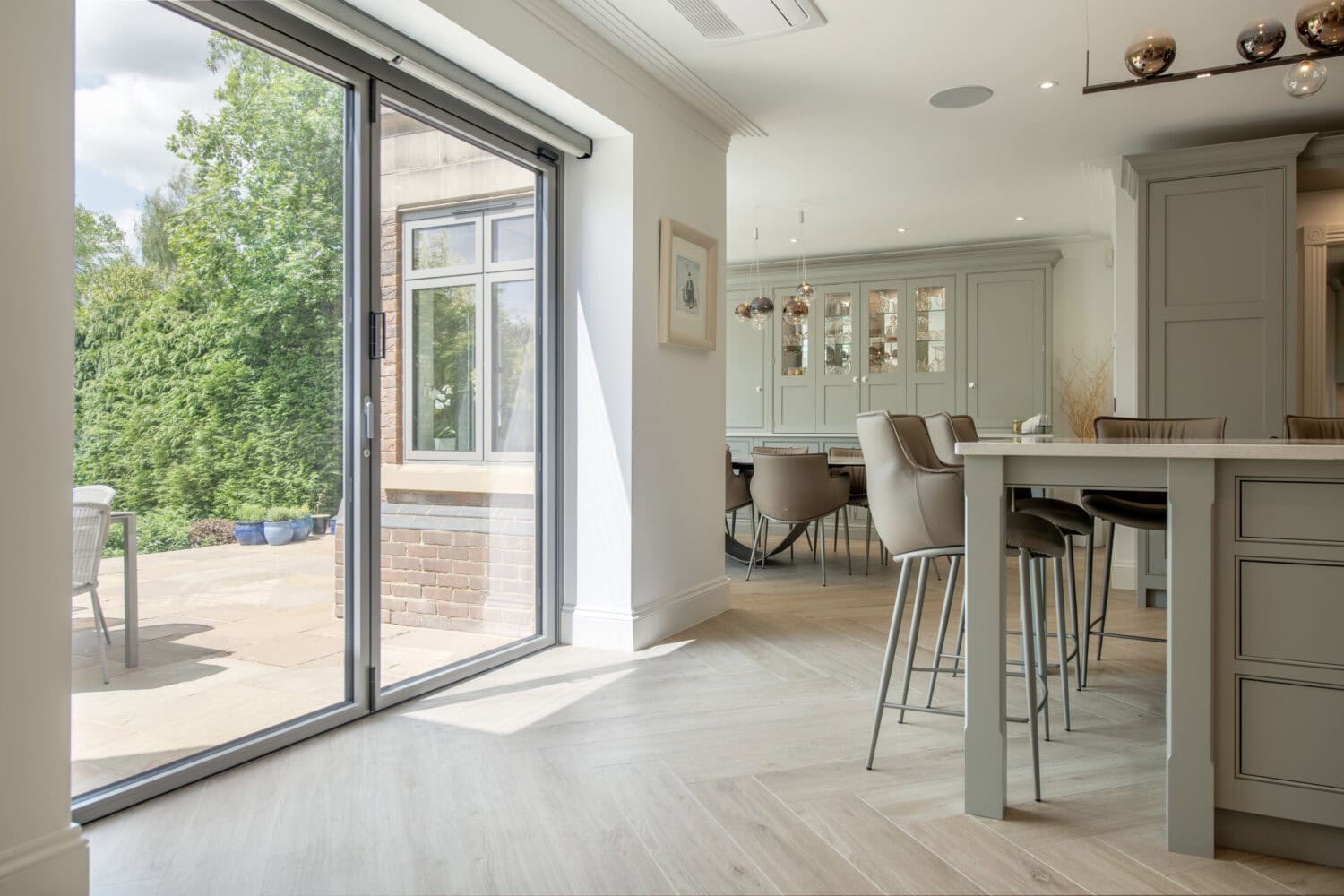 Modern kitchen and dining area with light wood floors, neutral cabinetry, and a large glass sliding door opening to a sunny patio with outdoor seating and lush greenery.