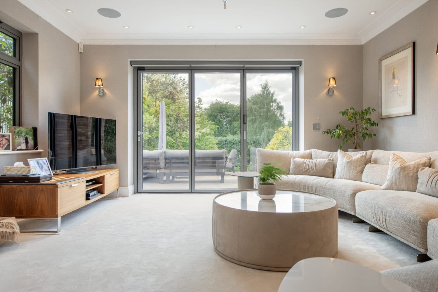 A modern living room with a curved beige sofa, round coffee table, large flat-screen TV, light walls, and large sliding glass doors opening to a patio with greenery outside.