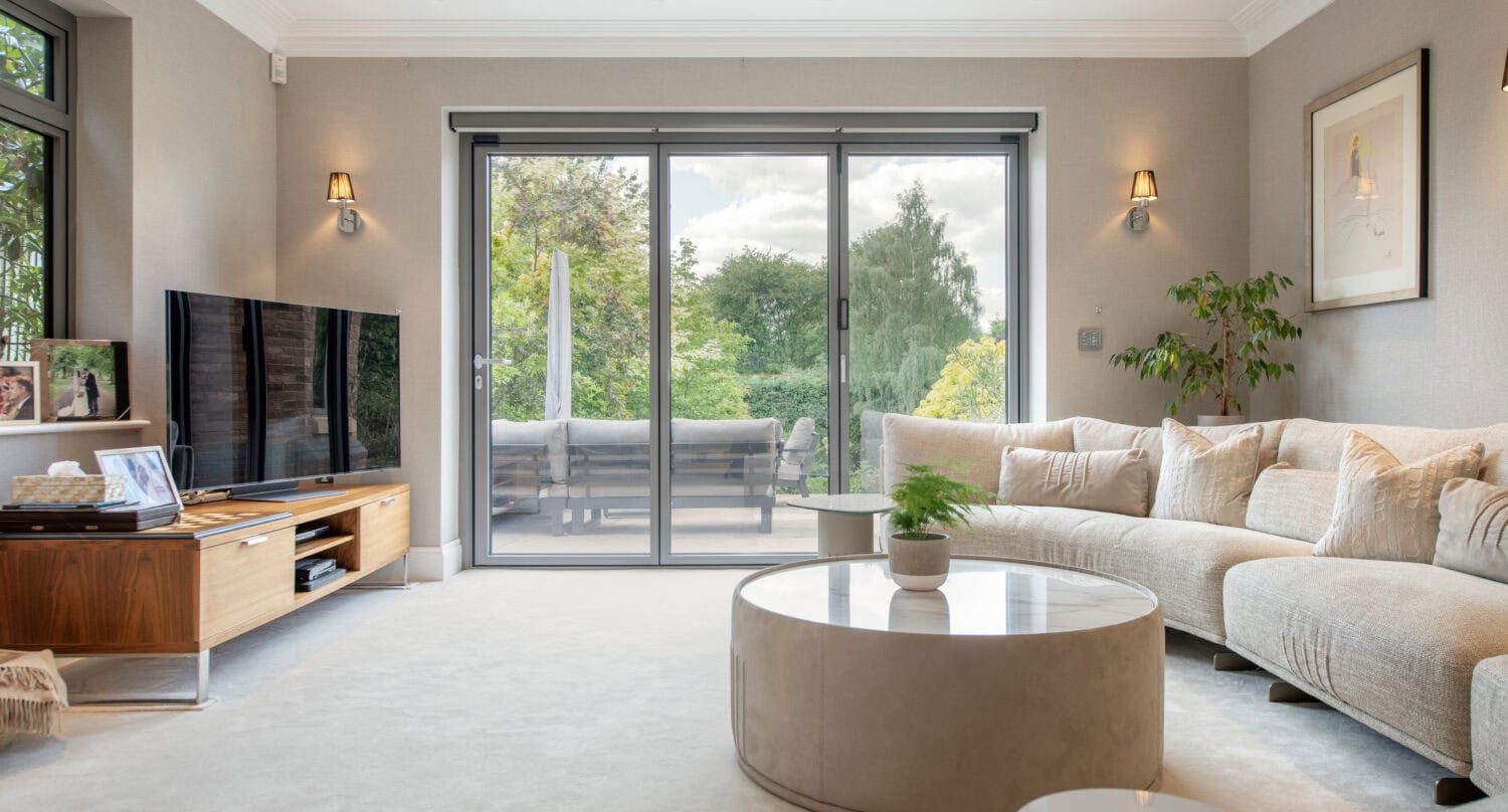 A modern living room with a curved beige sofa, round coffee table, large flat-screen TV, light walls, and large sliding glass doors opening to a patio with greenery outside.