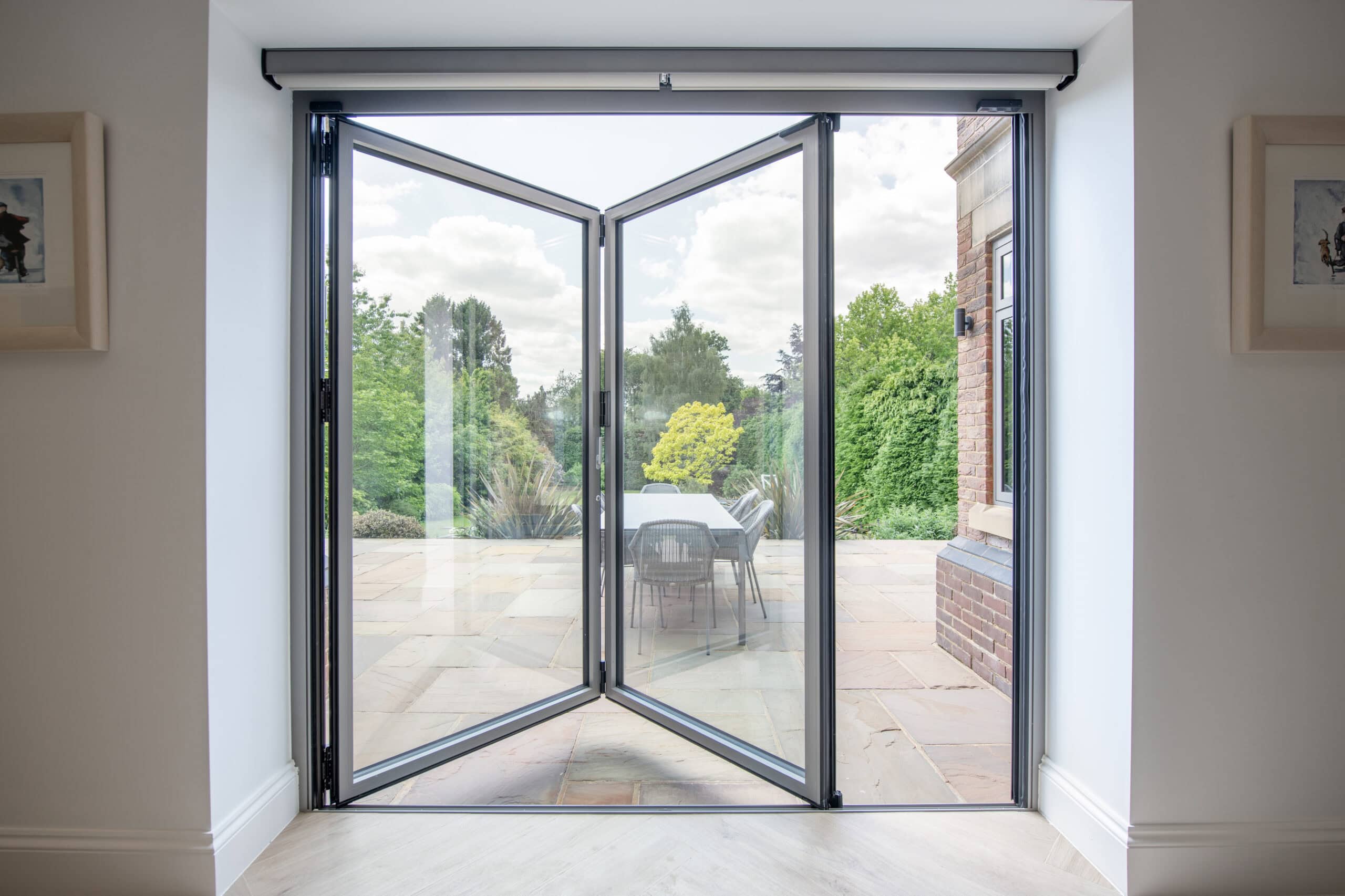 Open glass bifold doors leading to a patio with an outdoor table and chairs, surrounded by lush green trees and plants under a cloudy sky.