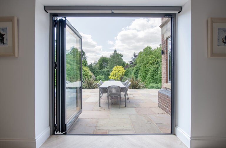 Open glass patio doors lead to a stone terrace with a table and chairs, surrounded by lush green trees and plants under a partly cloudy sky.