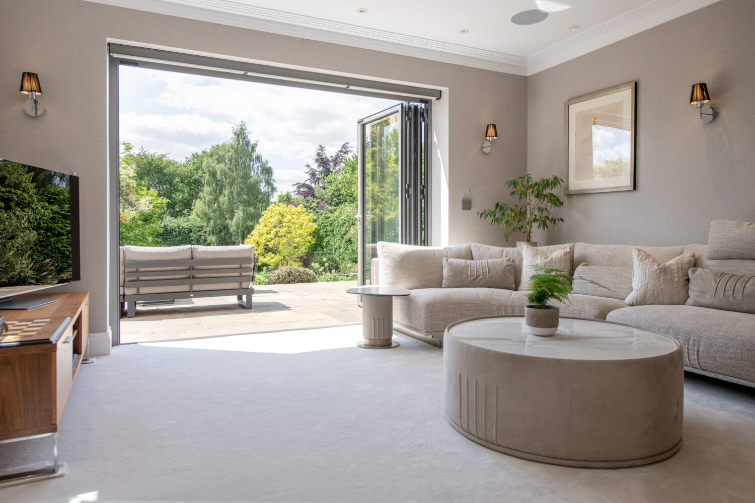 A bright, modern living room with a beige sectional sofa, round coffee table, and wall-mounted TV. Glass doors open to a patio with outdoor seating and a lush green garden view.