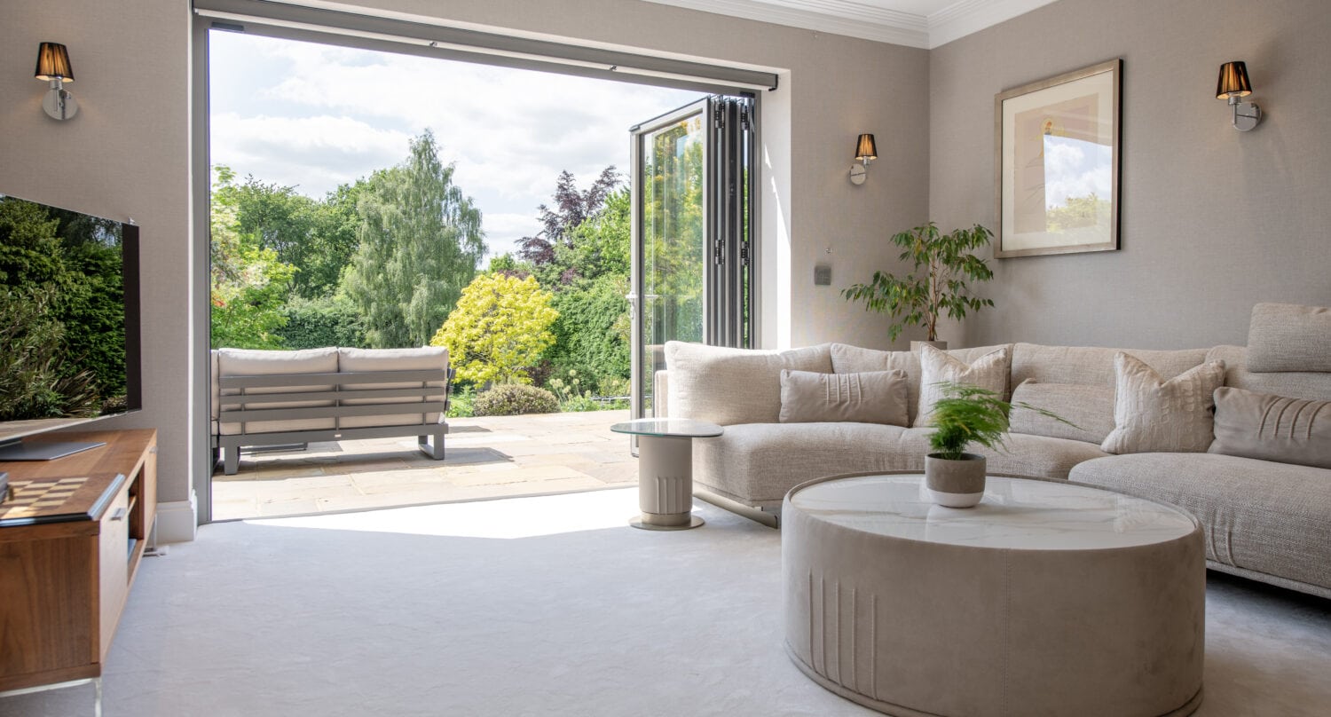 A bright, modern living room with a beige sectional sofa, round coffee table, and wall-mounted TV. Glass doors open to a patio with outdoor seating and a lush green garden view.