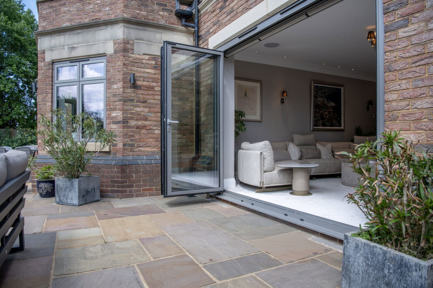 Modern brick house with large glass doors open to a stone patio, revealing a stylish living room with light-colored furniture and wall art. Potted plants decorate both the interior and exterior spaces.