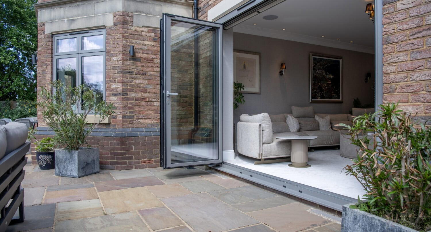 Modern brick house with large glass doors open to a stone patio, revealing a stylish living room with light-colored furniture and wall art. Potted plants decorate both the interior and exterior spaces.