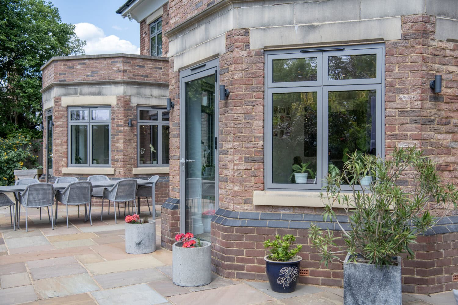 A modern brick house with large windows, a glass door, potted plants, and a spacious stone patio. There is an outdoor dining table with chairs and greenery in the background.