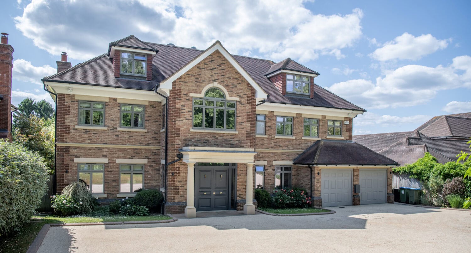 A large, modern brick house with three stories, arched windows, a double front door, and an attached two-car garage. The driveway is spacious, surrounded by greenery, under a partly cloudy sky.