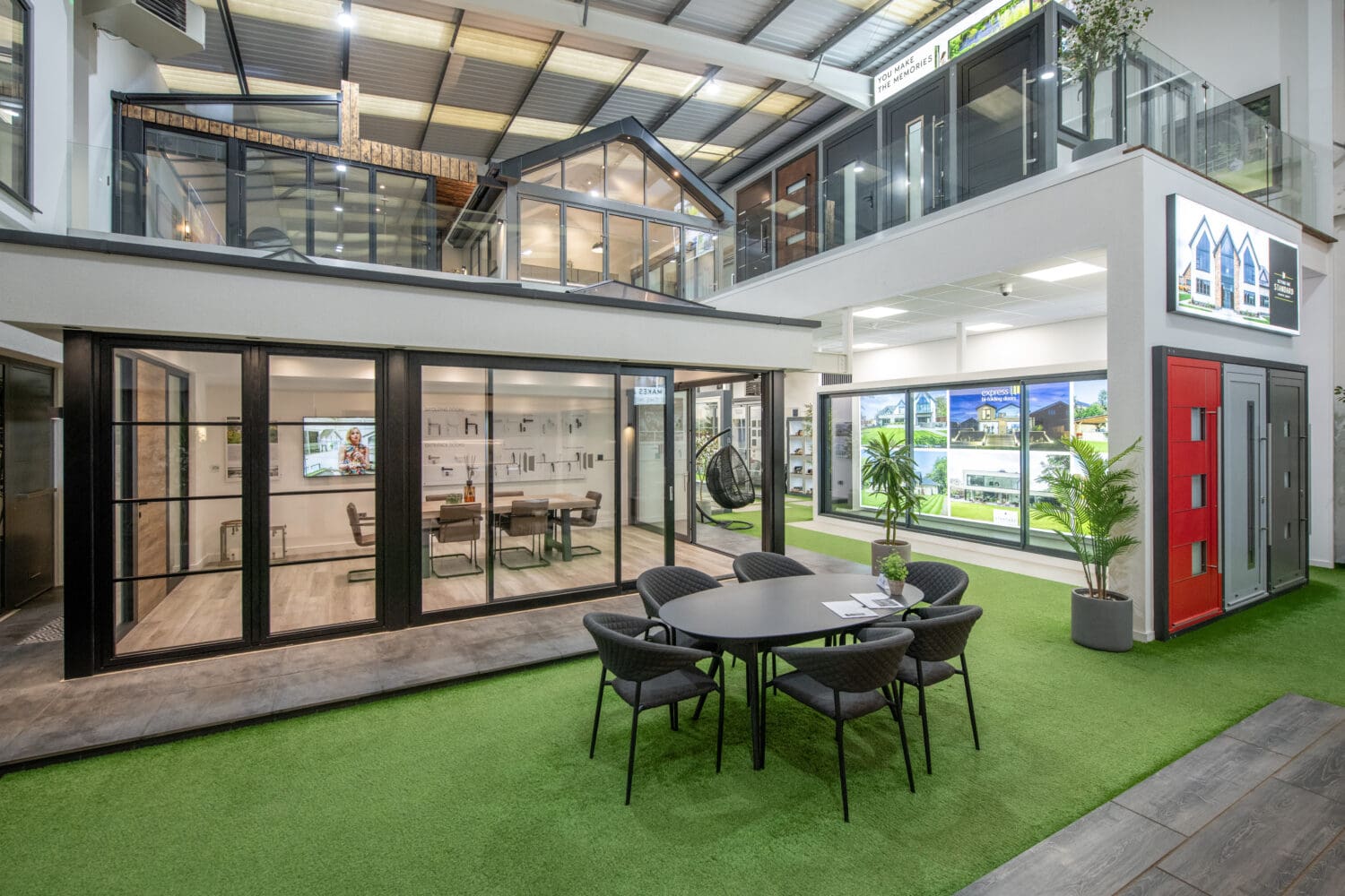 Modern showroom with large glass windows, a meeting table on artificial grass, and displays of bespoke glass solutions, front doors, and home designs. Potted plants add freshness to the bright upper level with stylish glass railings.