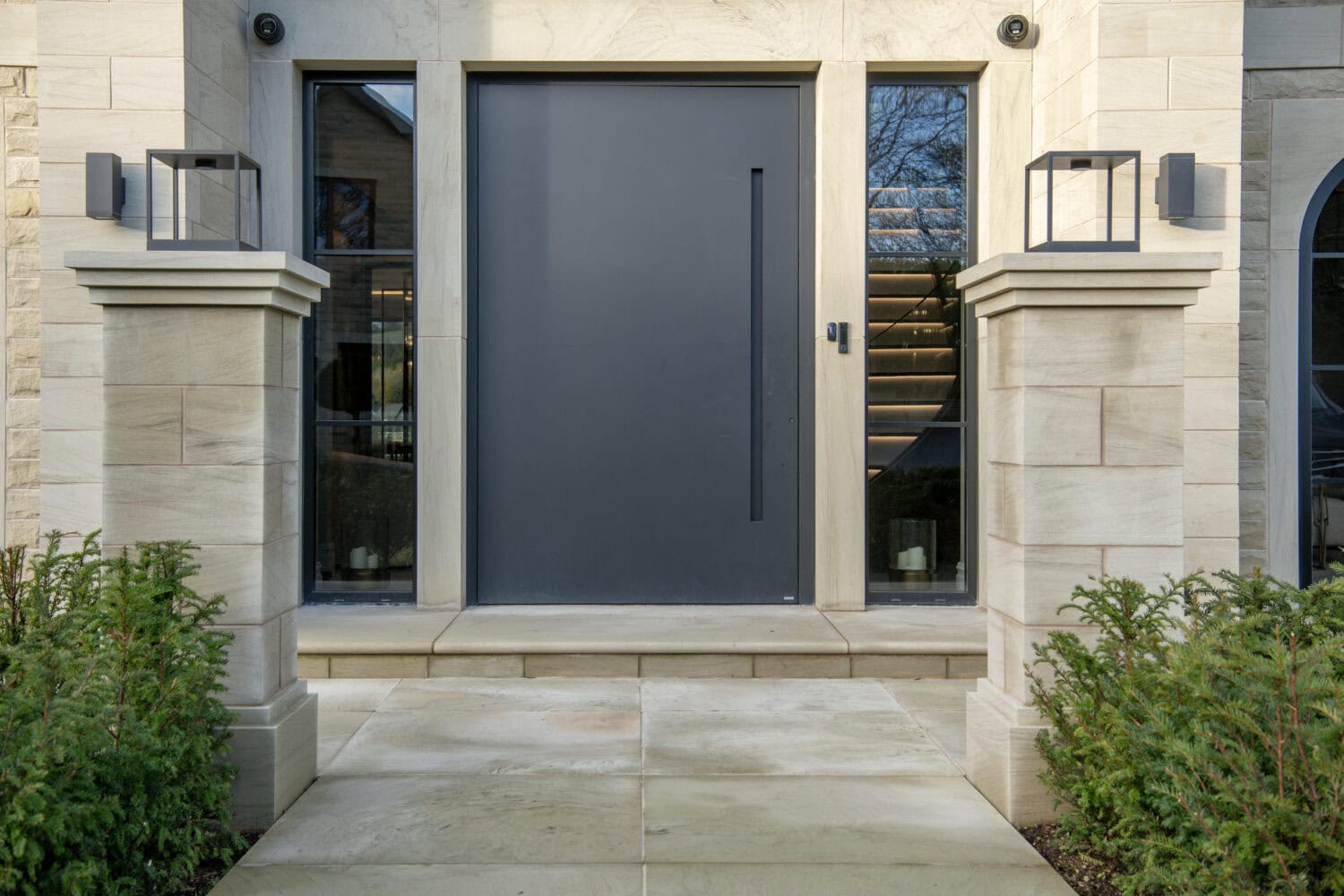 A sleek, modern pivot door in dark grey installed at a contemporary home entrance, flanked by large glass windows and stone pillars, enhancing curb appeal.