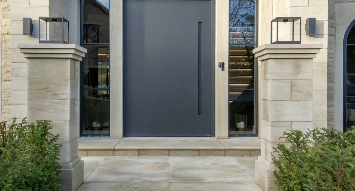A sleek, modern pivot door in dark grey installed at a contemporary home entrance, flanked by large glass windows and stone pillars, enhancing curb appeal.