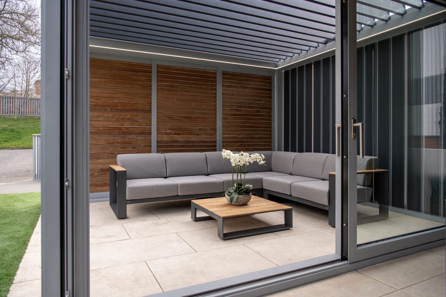 Elegant outdoor seating area with modern bi-folding doors, grey sofa, wooden coffee table, and stylish wooden wall panel, perfect for outdoor entertaining and seamless indoor-outdoor flow.