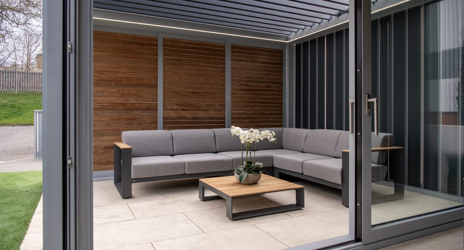 Elegant outdoor seating area with modern bi-folding doors, grey sofa, wooden coffee table, and stylish wooden wall panel, perfect for outdoor entertaining and seamless indoor-outdoor flow.