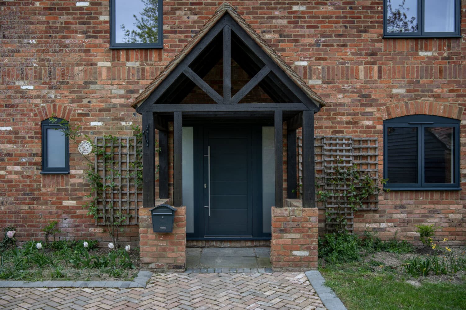 Stylish black front door with a wooden canopy, set in a brick house with garden plants and windows, ideal for enhancing home entrance appeal.