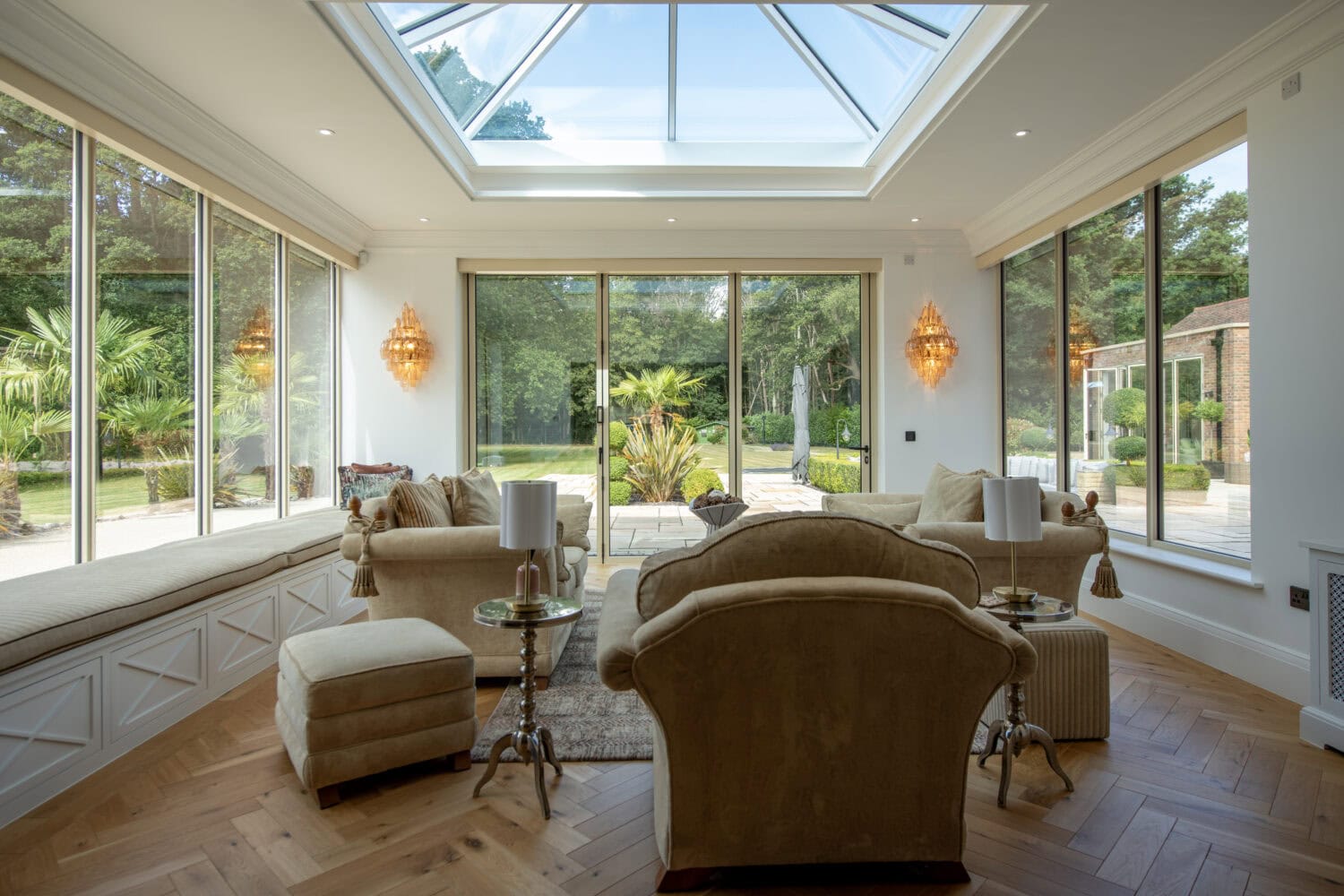 Spacious living room featuring large windows and a skylight, offering natural light and views of a lush garden, with comfortable seating and elegant decor.