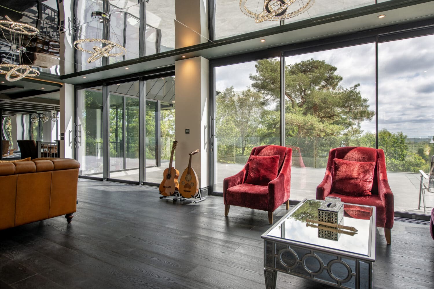 Stylish living space featuring large bi-folding glass doors opening to scenic outdoor views, with red armchairs, a glass coffee table, and musical instruments.