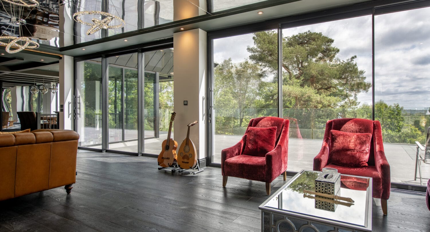 Stylish living space featuring large bi-folding glass doors opening to scenic outdoor views, with red armchairs, a glass coffee table, and musical instruments.