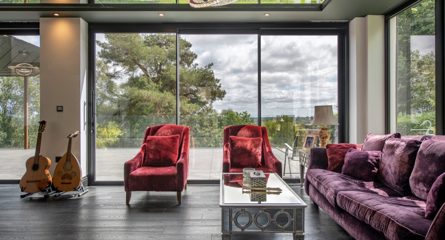 Modern living room with large glass bi-folding doors opening to outdoor greenery, featuring plush purple and red seating, and a sleek coffee table.