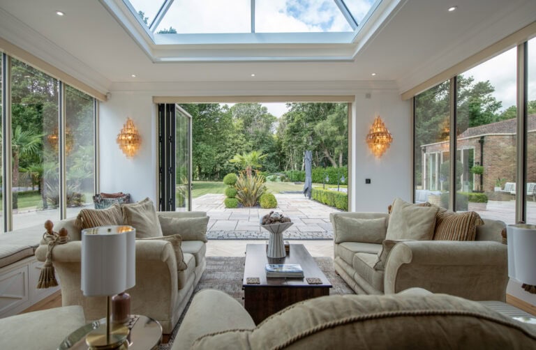 Elegant living room with large glass bi-fold doors opening to a lush garden, featuring comfortable sofas and a skylight ceiling.