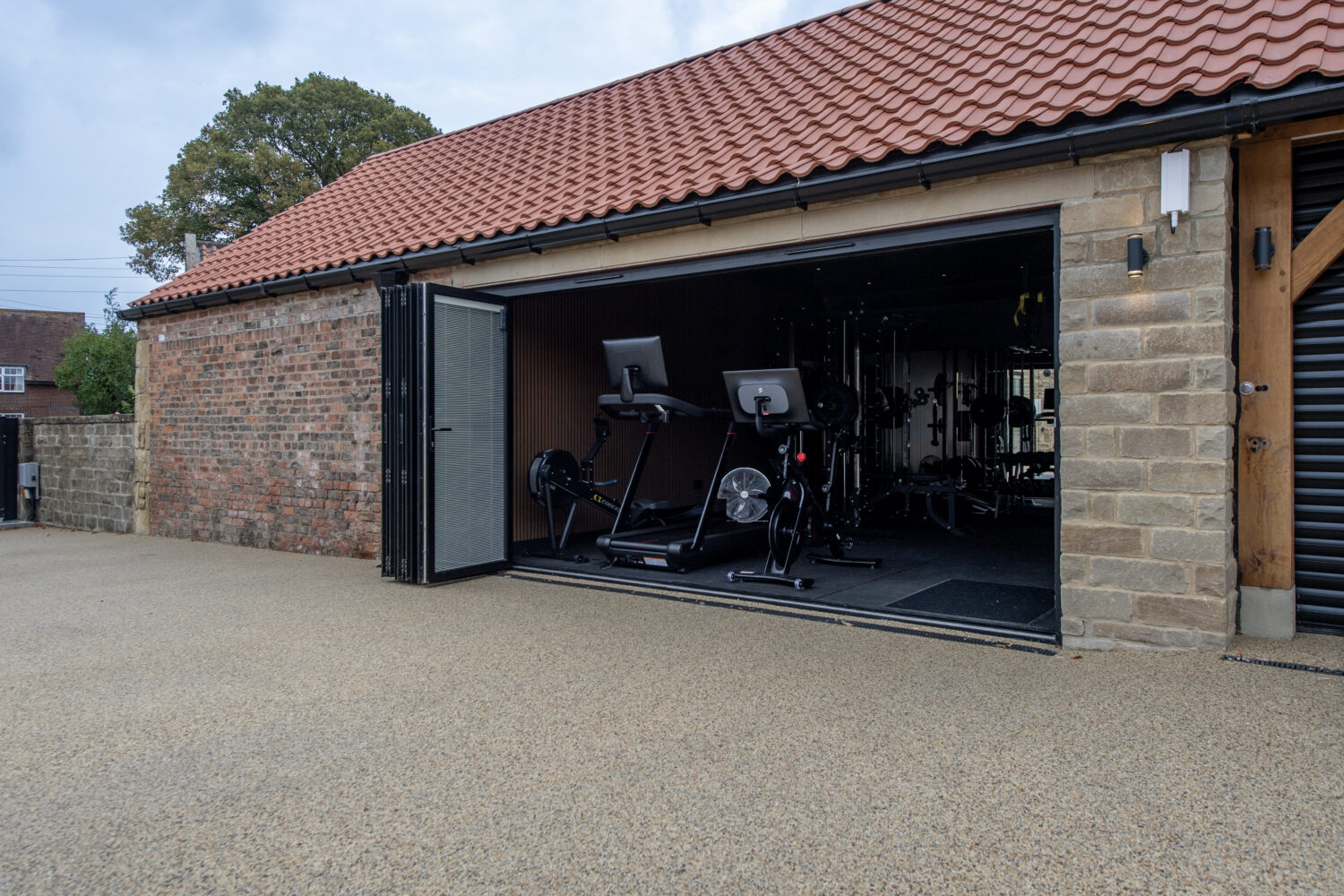 Modern garage with black bi-folding doors open to reveal gym equipment inside, set against a brick wall and red tiled roof, under a clear sky.