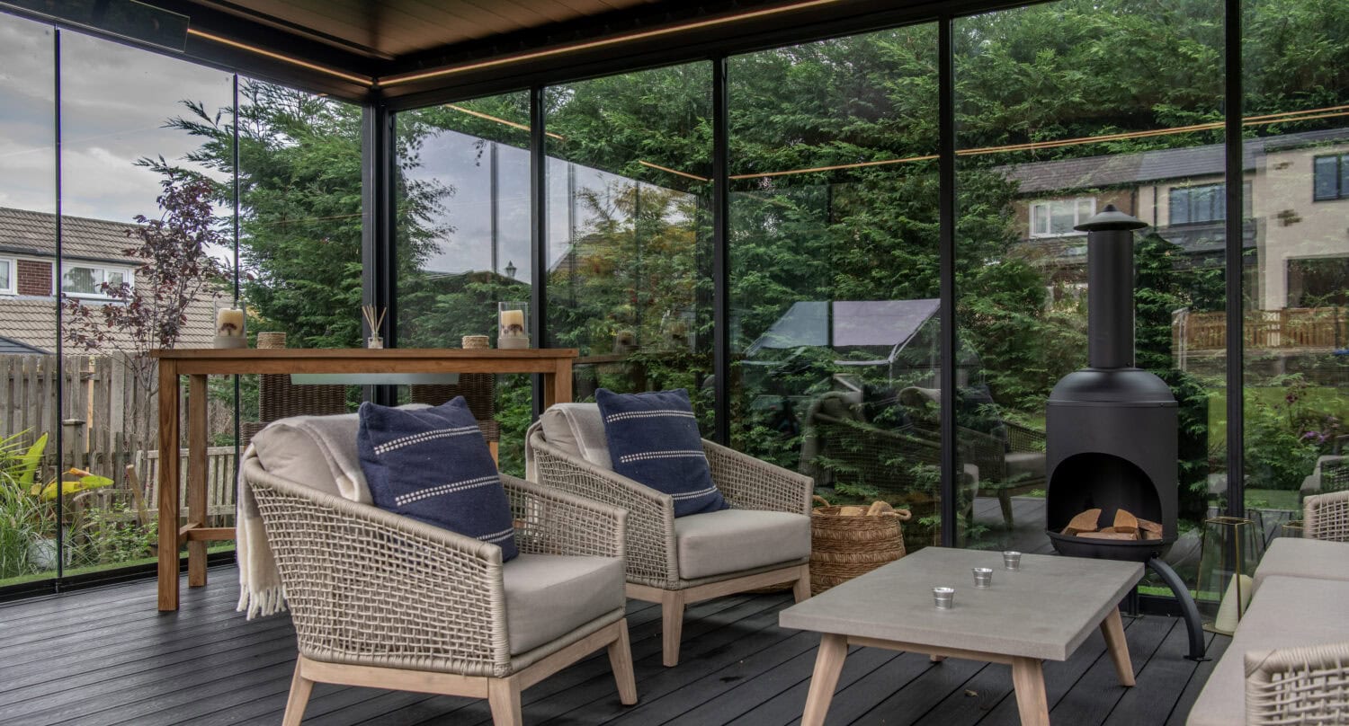 Contemporary enclosed patio featuring large glass bi-fold doors, wicker chairs with cushions, a fire pit, and a scenic garden view, perfect for outdoor living and solar shading.