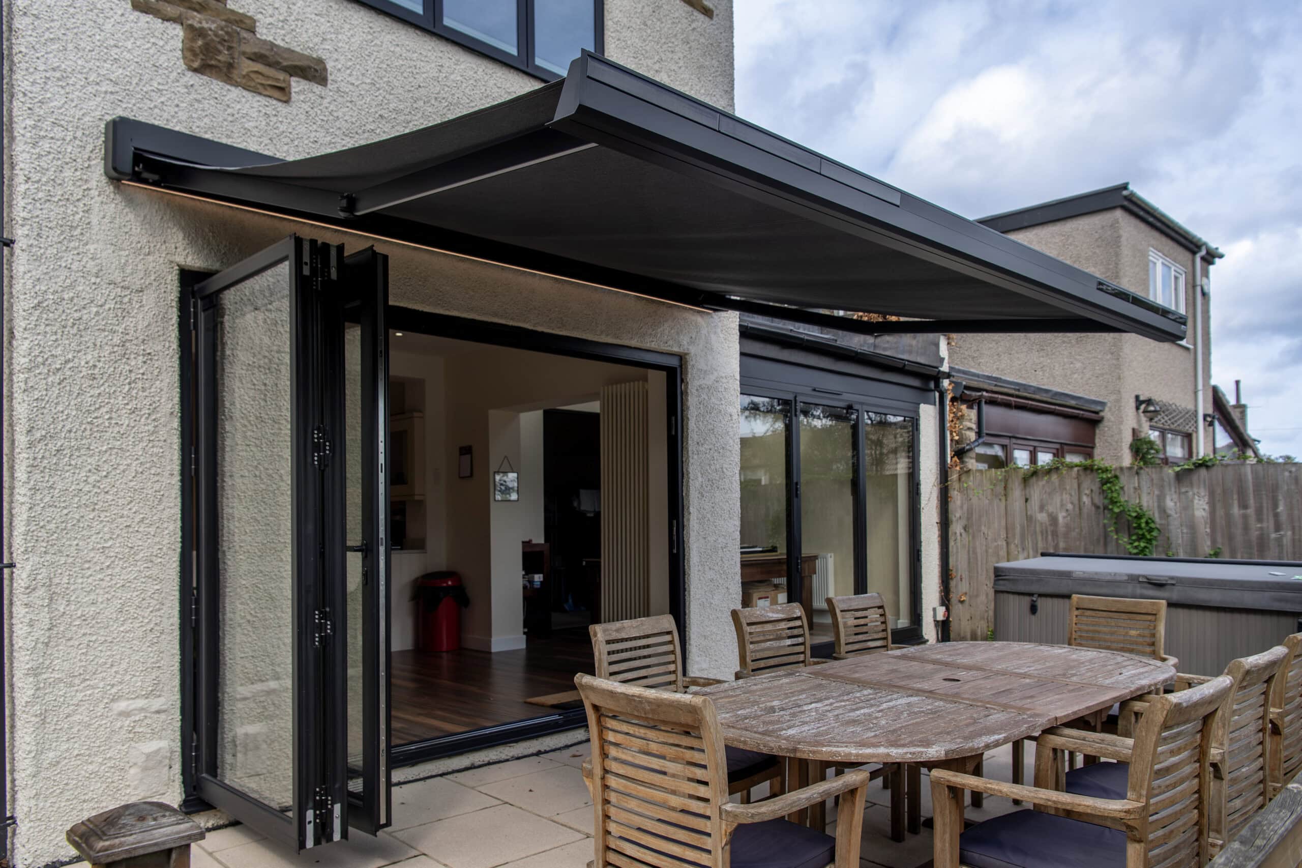 Modern black bi-folding doors opening to an outdoor patio with wooden dining table and chairs, under a retractable awning, in a residential setting.
